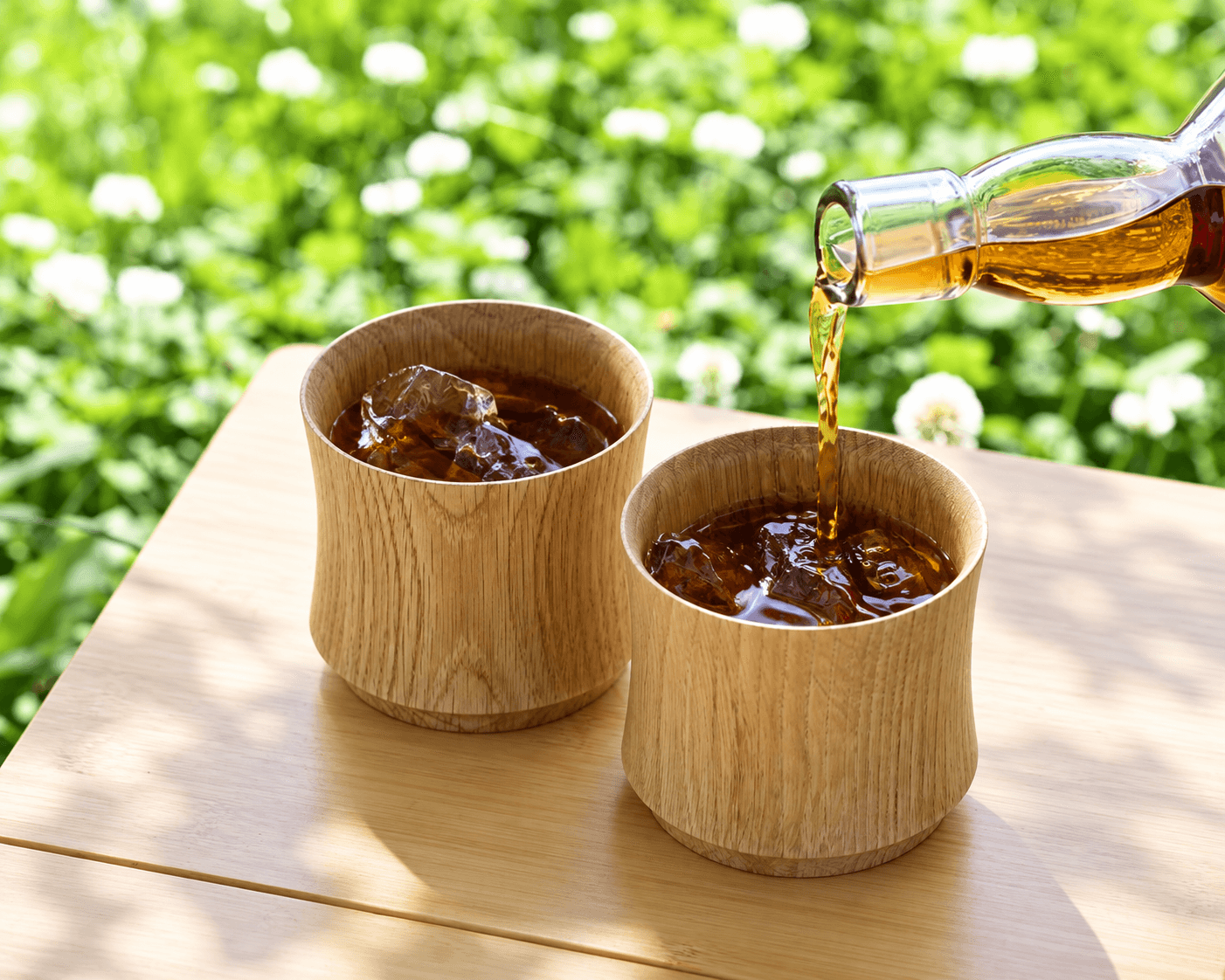 緑の庭を背景に、木の杯へ冷たい飲み物を注ぐ様子